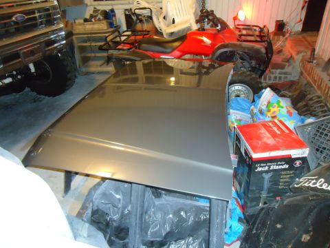 1986 Ford Bronco - A silver vehicle hood rests on a stand in a garage, with a red ATV and a portion of a Ford truck visible in the background. Several boxes,…