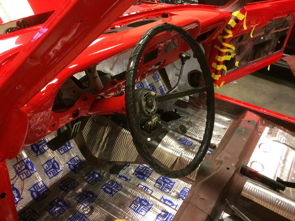 Interior of a red classic car with sound deadening on the floor and a black steering wheel.