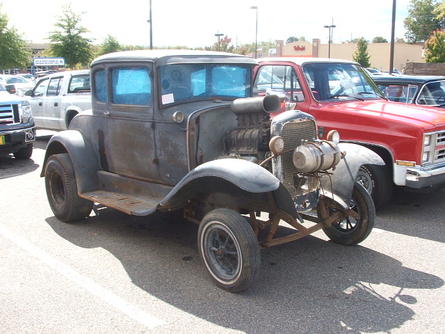 Weathered hot rod coupe with supercharger and blue windows parked next to a red pickup truck.