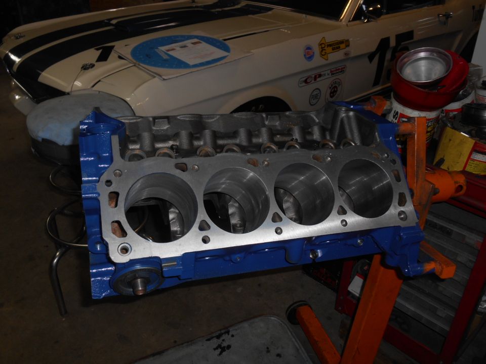 Blue V8 engine block on an orange stand, with a classic white car with blue stripes in the background.