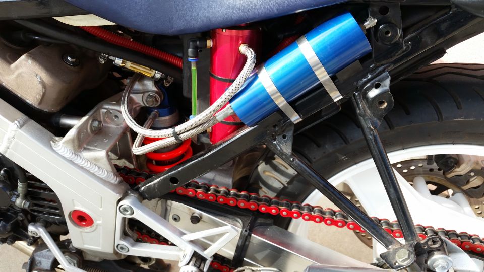 Close-up of a motorcycle's rear suspension with blue reservoir shock and red chain.