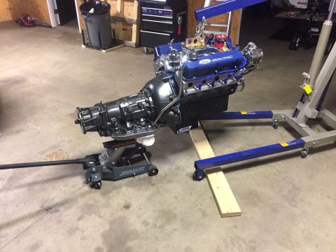 1982 Ford Bronco - A Ford Racing engine and transmission assembly is suspended by an engine hoist and supported by a floor jack in a garage setting.