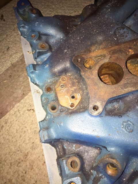 1978 Ford Bronco - A close-up view of a blue painted engine intake manifold with visible rust and corrosion. The manifold has several bolt holes and ports, wi…