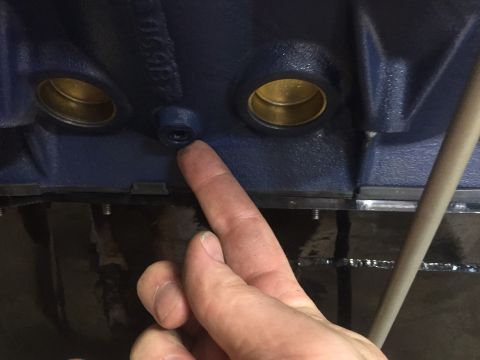 1982 Ford Bronco - A finger points to a small threaded hole on the side of a dark blue engine block. Two brass-colored bearing holes are visible to the left o…