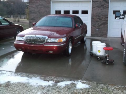 1999 Mercury Grand Marquis - A red Mercury Grand Marquis is parked on a wet driveway in front of a brick garage with white doors. Two white buckets and a red stool are…