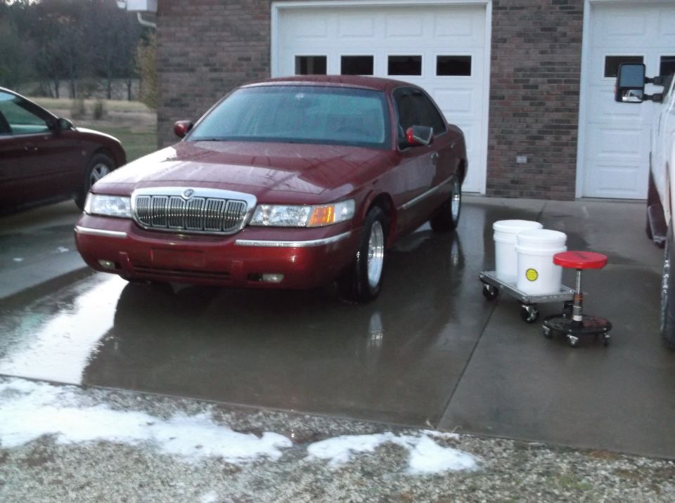 Red Mercury Grand Marquis parked on a wet driveway in front of a garage with two buckets and a stool nearby.