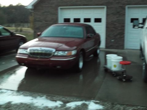 1999 Mercury Grand Marquis - A maroon sedan is parked on a wet driveway in front of a brick garage with two white doors. Two white buckets and a red stool are on a roll…