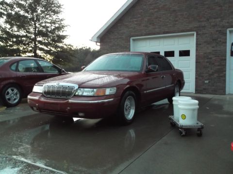 1999 Mercury Grand Marquis - A maroon sedan is parked in front of a brick garage with a white door. Two white buckets on a rolling cart are visible next to the car.