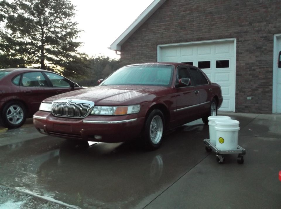 Maroon sedan parked in front of a brick garage with two white buckets on a cart.
