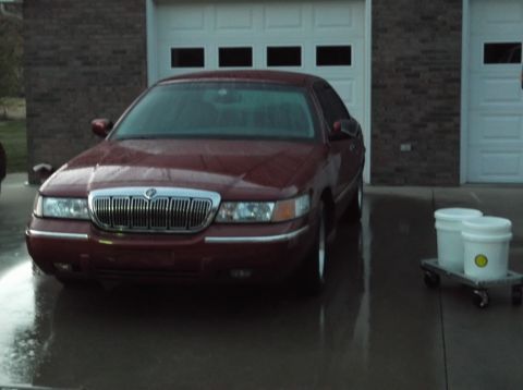 1999 Mercury Grand Marquis - A maroon 1999 Mercury Grand Marquis is parked in front of a garage with a wet driveway. Two white buckets on a dolly are visible to the rig…