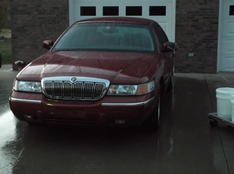 1999 Mercury Grand Marquis - A maroon Mercury Grand Marquis is parked in front of a brick garage with white doors. The car appears wet, possibly from rain, with water d…