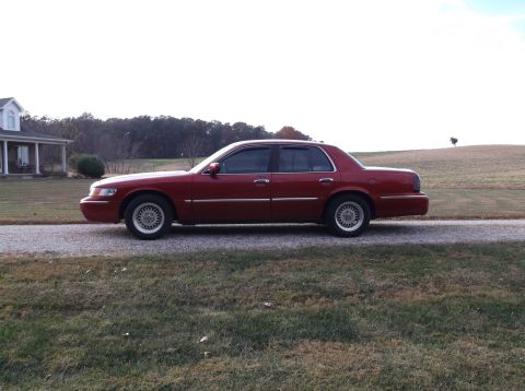 1999 Mercury Grand Marquis - A side profile of a maroon 1999 Mercury Grand Marquis sedan is parked on a gravel driveway in front of a house and a wooded area. The car h…