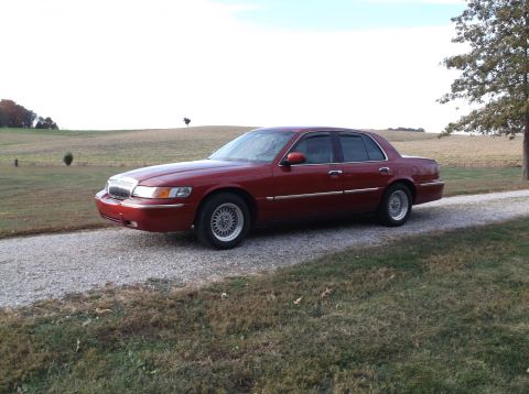1999 Mercury Grand Marquis - A red 1999 Mercury Grand Marquis sedan is parked on a gravel driveway with a grassy field and trees in the background.