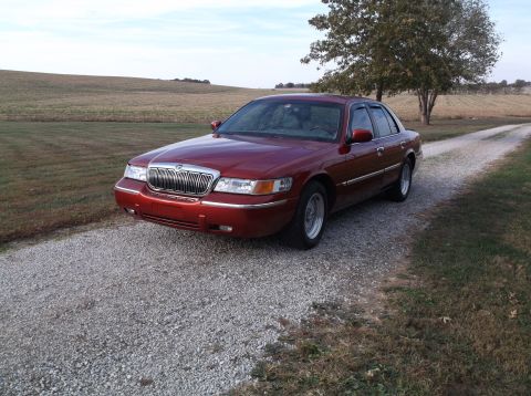 1999 Mercury Grand Marquis - A red 1999 Mercury Grand Marquis is parked on a gravel driveway next to a grassy field and a dirt road. The car is facing the front-left, w…