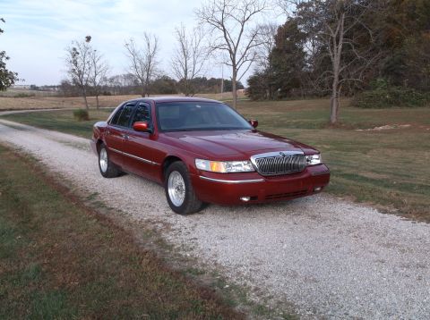1999 Mercury Grand Marquis - A red Mercury Grand Marquis sedan is parked on a gravel driveway in a rural setting with trees and fields in the background.