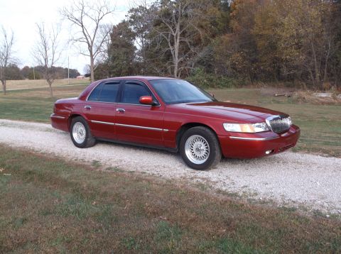1999 Mercury Grand Marquis - A dark red 1999 Mercury Grand Marquis sedan is parked on a gravel driveway with grass and trees in the background. The car is viewed from t…