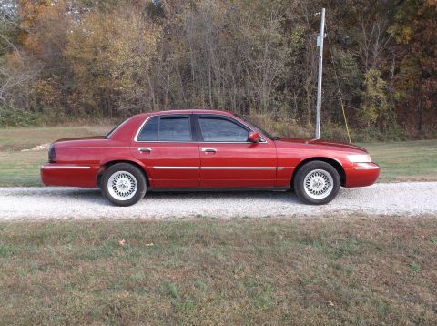 1999 Mercury Grand Marquis - A red 1999 Mercury Grand Marquis sedan is parked on a gravel driveway with a wooded area in the background. The car is viewed from the side…