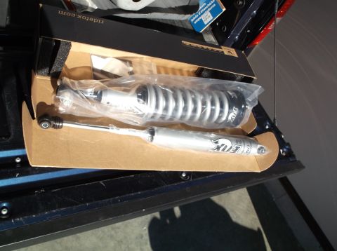 2005 Ford F-150 - A pair of new Fox 2.0 shocks are displayed in their original packaging inside the bed of a dark blue pickup truck. The shocks are silver wi…