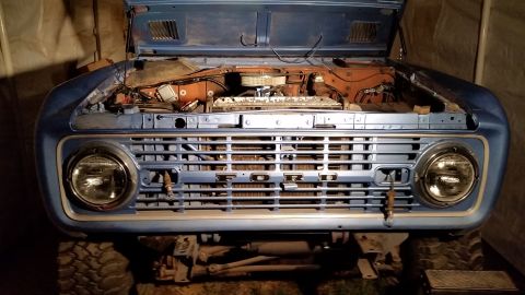 1973 Ford Bronco - Front view of a blue 1973 Ford Bronco with its hood open, revealing the engine bay. The vehicle has a prominent silver grille with the "FOR…