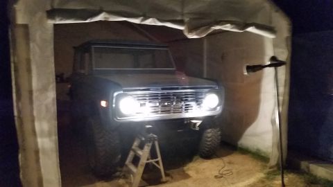 1973 Ford Bronco - A Ford Bronco is seen from the front, parked inside a temporary structure with its headlights illuminated.