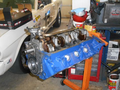 1972 Ford Bronco - A blue engine block with internal components and timing chain sits on an orange engine stand, with a white car in the background.
