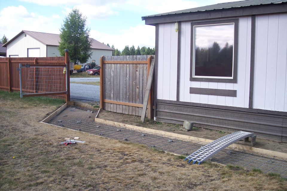 Two vehicles, a red SUV and a yellow truck, parked on gravel near a fence and a mobile home