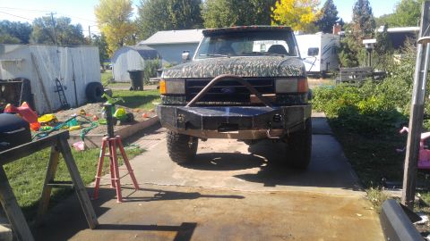1991 Ford Bronco - A modified Ford Bronco with a camouflage hood and a custom heavy-duty bumper sits in a driveway with various yard items and sheds in the ba…
