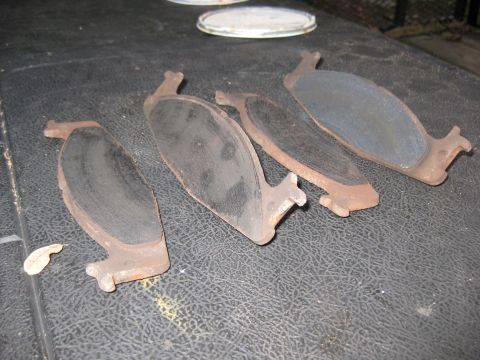 1994 Ford Bronco - Four used brake pads are laid out on a textured black surface. The brake pads show signs of wear and rust on their metal backing plates.