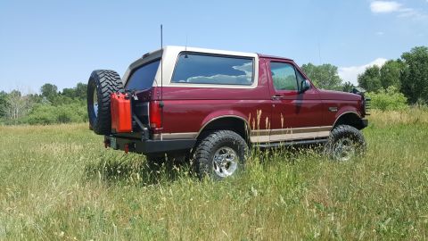 1996 Ford Bronco - A red 1996 Ford Bronco with a tan roof is parked in a grassy field under a clear blue sky. The vehicle features a spare tire and a red jerr…
