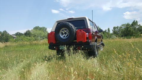 1996 Ford Bronco - The rear of a maroon and tan Ford Bronco is parked in a grassy field under a blue sky with scattered clouds. It features a spare tire mount…