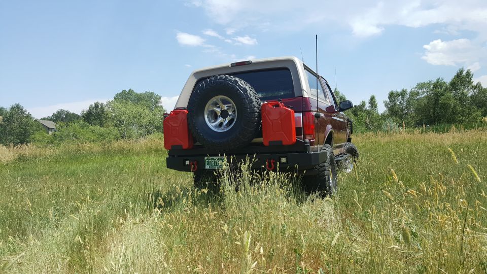 Rear view of a maroon and tan Ford Bronco with a spare tire, jerry cans, and custom bumper in a grassy field.