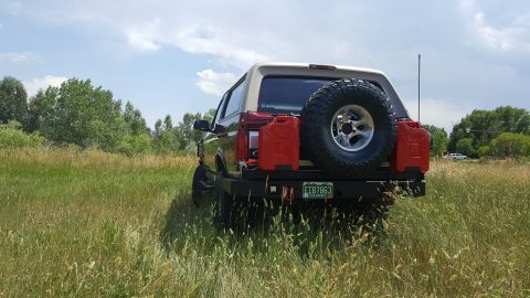 1996 Ford Bronco - The rear view of a lifted, dark red 1996 Ford Bronco parked in a grassy field. It features a spare tire mounted on the rear, two red jerry…