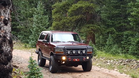 1996 Ford Bronco - A maroon 1996 Ford Bronco is parked on a dirt road in a forest. The SUV features an aftermarket front bumper with auxiliary lights and red…