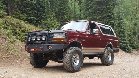 1996 Ford Bronco - A lifted, maroon 1996 Ford Bronco with a custom front bumper and brush guard is parked on a dirt road in a forest.