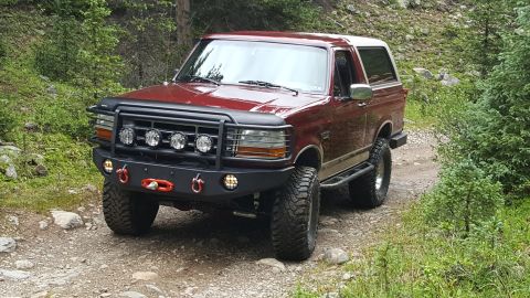 1996 Ford Bronco - A maroon 1996 Ford Bronco with an aftermarket front bumper and brush guard is parked on a dirt road in a wooded area. The vehicle has large…