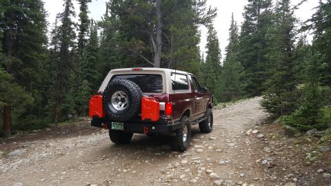 1996 Ford Bronco - A maroon 1996 Ford Bronco with a tan top is parked on a gravel road in a forest. The vehicle has a spare tire mounted on the back, two red…