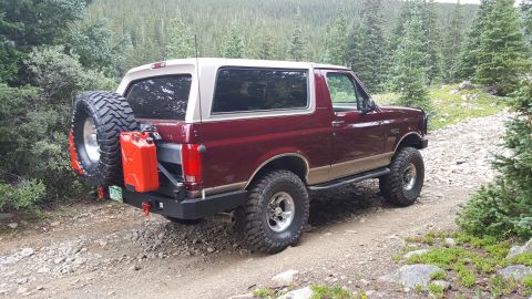 1996 Ford Bronco - The rear passenger side of a maroon and tan 1996 Ford Bronco is shown parked on a rocky dirt road in a forest. The vehicle features a spare…