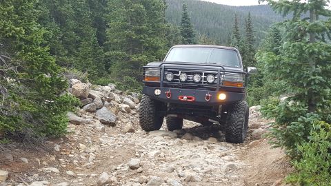 1996 Ford Bronco - A dark-colored Ford Bronco is navigating a rocky, mountainous trail surrounded by pine trees. The off-road vehicle features a custom bumper…