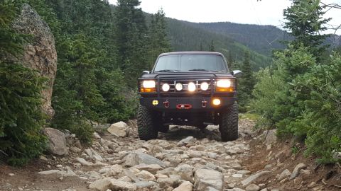 1996 Ford Bronco - A dark colored Ford Bronco is parked on a rocky, mountainous trail surrounded by pine trees. The vehicle has a custom bumper with multiple…