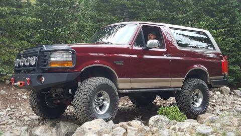 1996 Ford Bronco - A maroon 1996 Ford Bronco with a tan lower body accent is parked on a rocky, wooded trail. The SUV features an aftermarket front bumper wit…