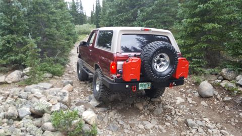 1996 Ford Bronco - A maroon 2-door SUV with a tan top is driving on a rocky, wooded trail. The vehicle has a spare tire mounted on the back, two red jerry can…
