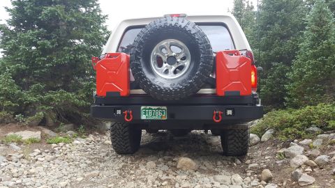 1996 Ford Bronco - The rear of a white 1996 Ford Bronco is shown parked on a rocky, wooded trail. It is equipped with a black aftermarket bumper, two red jerr…