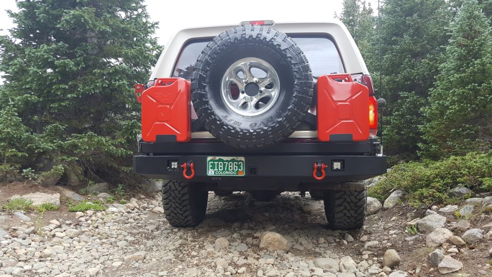 Rear view of a white 1996 Ford Bronco with a spare tire, jerry cans, and aftermarket bumper on a rocky trail.