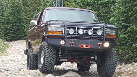 1996 Ford Bronco - A lifted, dark red 1996 Ford Bronco with an aftermarket bumper and KC lights is parked on a rocky trail surrounded by trees. The front of t…