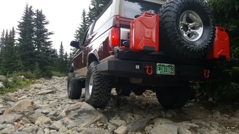 1996 Ford Bronco - The rear of a maroon Ford Bronco is shown driving on a rocky mountain trail. It features a spare tire mounted on the back, two red jerry ca…