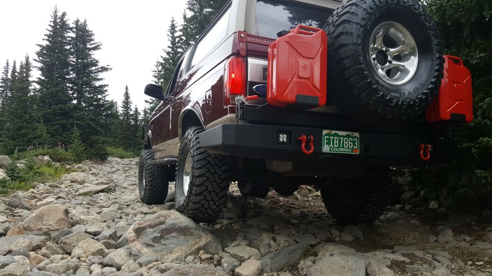 Rear view of a maroon Ford Bronco on a rocky trail with a spare tire and jerry cans.