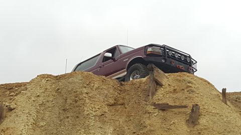 1996 Ford Bronco - A maroon 1996 Ford Bronco is shown from a low angle, climbing a steep dirt mound with visible tire tracks. The vehicle has a black aftermar…