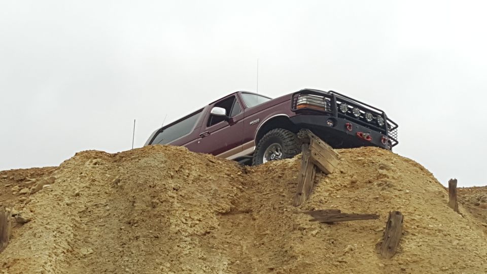 Maroon 1996 Ford Bronco climbing a dirt mound with aftermarket bumper and lights.
