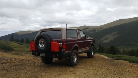 1996 Ford Bronco - The rear driver's side of a maroon and tan 1996 Ford Bronco is parked on a dirt road with a mountainous, forested landscape in the backgrou…