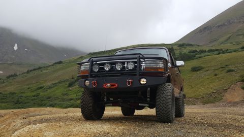 1996 Ford Bronco - A modified maroon Ford Bronco is parked on a dirt road in a mountainous, cloudy landscape. The vehicle features a large aftermarket bumper…